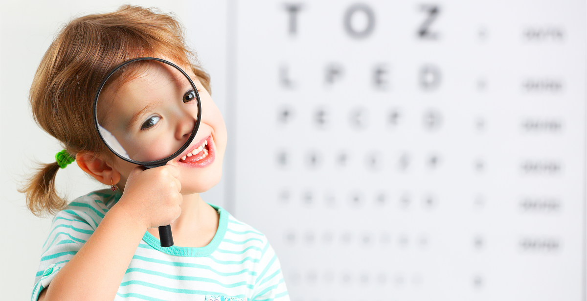 ילדה קטנה עם קוקיות מסתכלת מבעד לזכוכית מגדלת, מאחוריה לוח קריאה של בדיקות ראיה