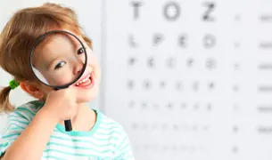 ילדה קטנה עם קוקיות מסתכלת מבעד לזכוכית מגדלת, מאחוריה לוח קריאה של בדיקות ראיה