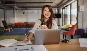 A woman holding a cup of coffee works in the office and has a laptop next to her