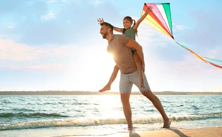 אבא מרים את הבת שלו על  הגב והילדה מחזיקה עפיפון בים