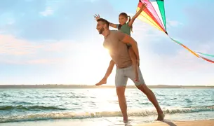 אבא מרים את הבת שלו על הגב והילדה מחזיקה עפיפון בים