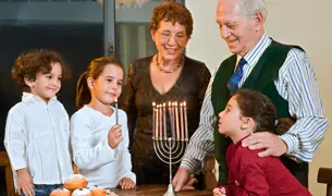 סבא וסבתא מדליקים נרות חנוכה ביחד עם הנכדים, על השולחן יש חנוכיה וסופגניות