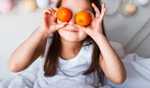 ילדה יושבת על המיטה ומחזיקה שתי קלמנטינות ליד העיניים