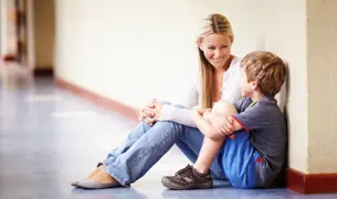 מורה ותלמיד צעיר יושבים במסדרון בבית הספר והיא מנחמת אותו